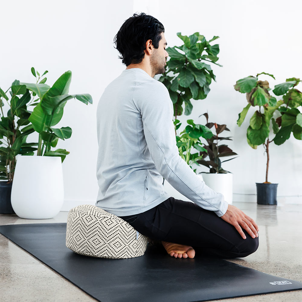 The Calm - Meditation Cushion in beige and black colour - Modern City Day - round modern cushion with removable washable cover photographed in use with fit man seated and meditating with beautiful plants in the background.