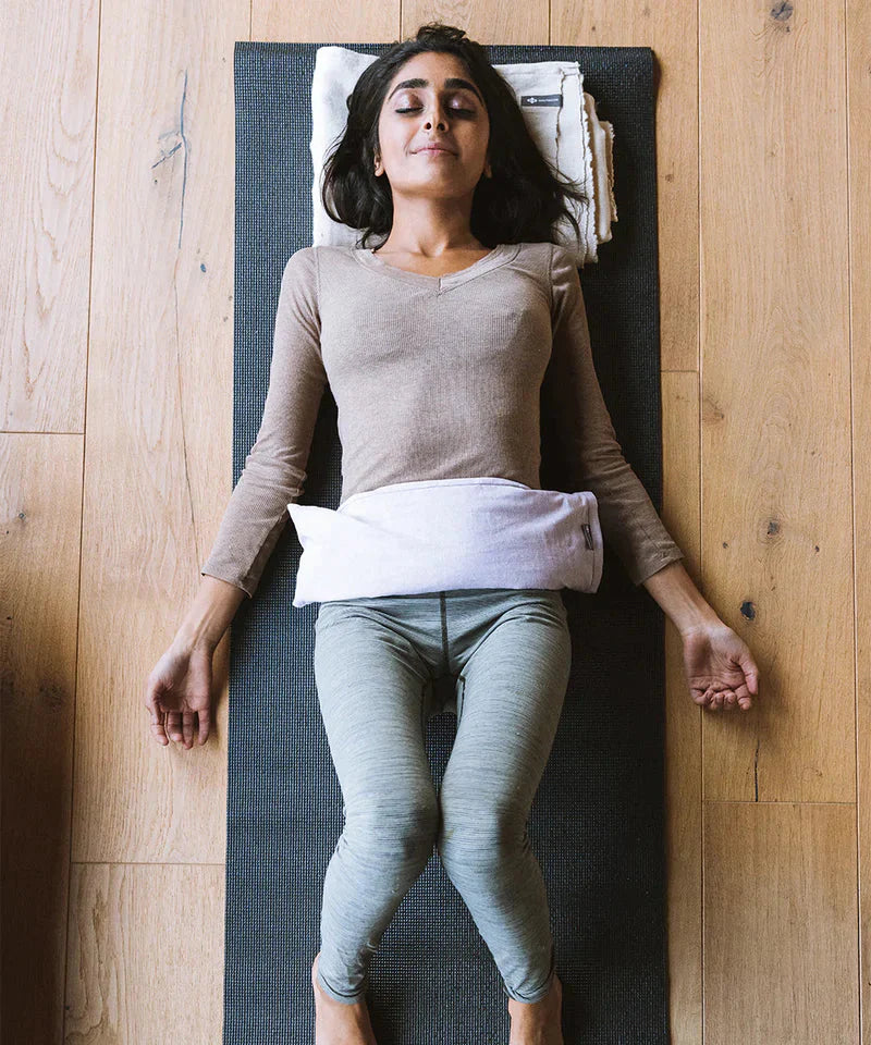 Photograph of woman in yoga nidra pose with Iris coloured Linen Hot + Cold Therapy Pillow on her lower abdomen.