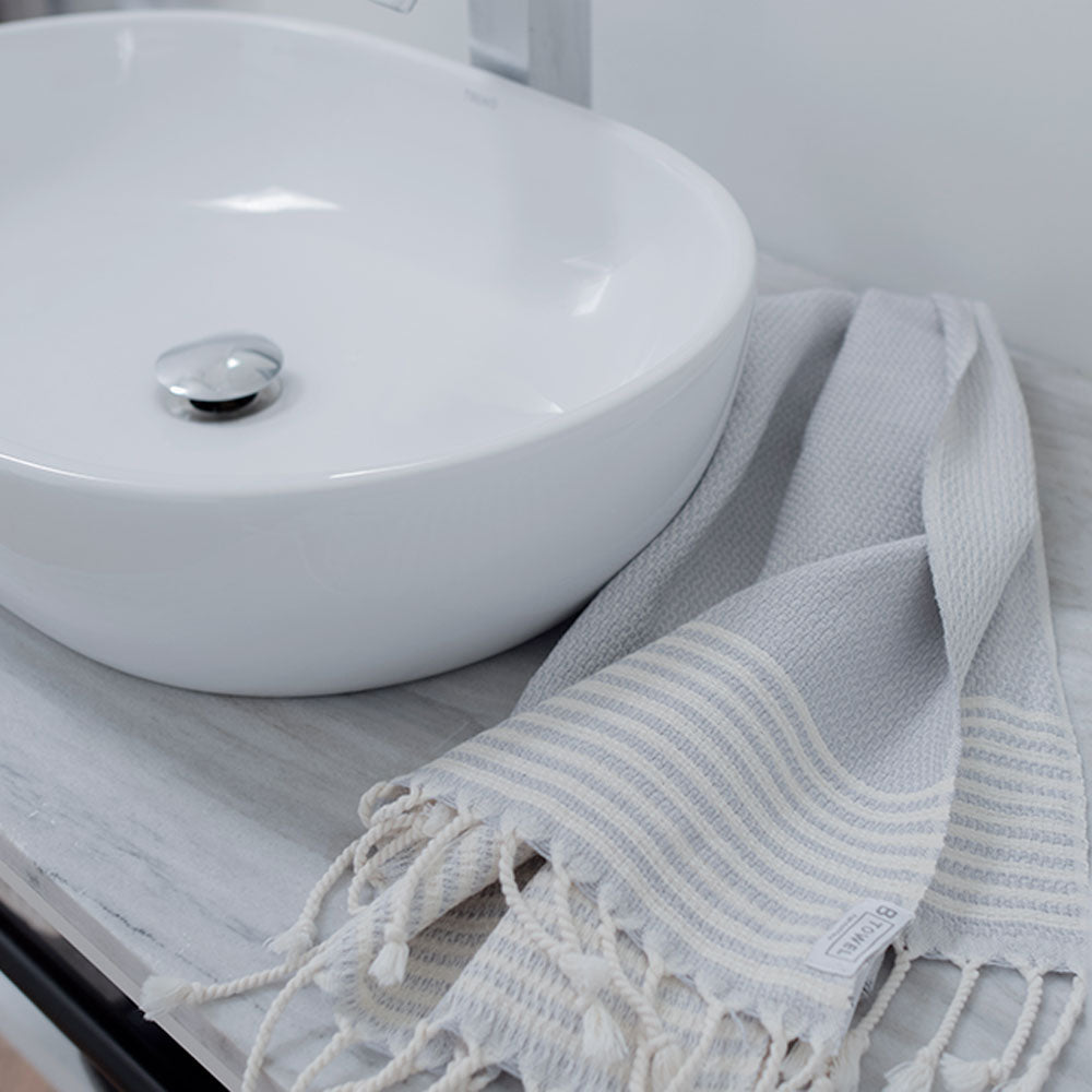 Lifestyle shot - Stone Grey - hand and face towel by B Yoga - Turkish Cotton laying beside tap of a white sink