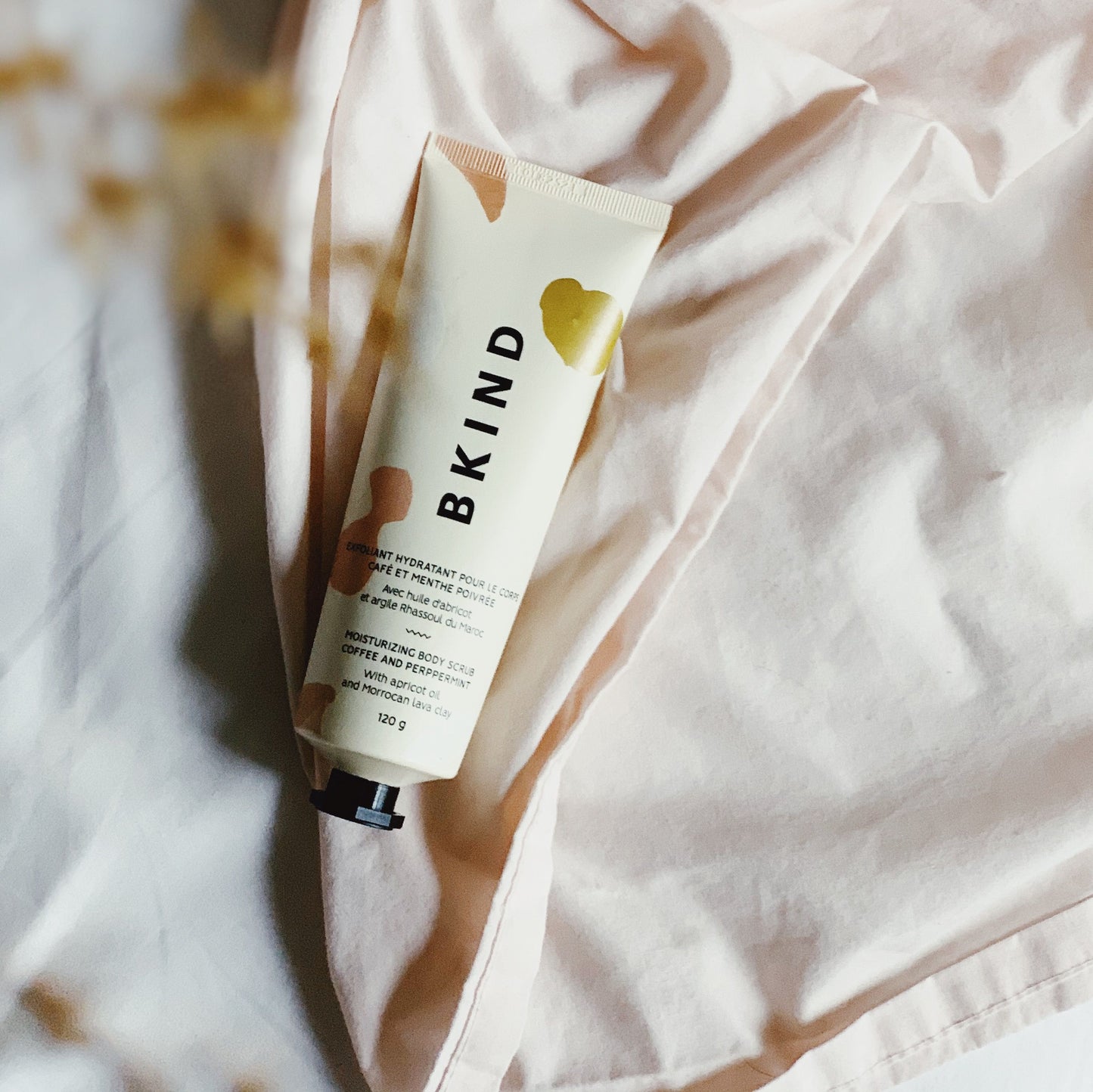 BKIND coffee and peppermint moisturizing scrub in a beige tube with peach designs on the tube displayed on a cloth background in dim lighting..