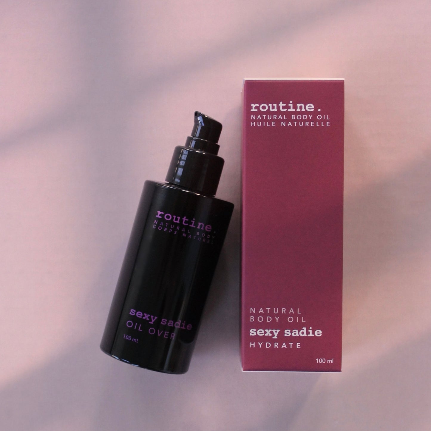 Magenta Box labelled ROUTINE body oil serum - SEXY SADIE - hydrate beside a black sleek bottle with magenta printing on it. Pump top included on the bottle. Light purple shadowed background.