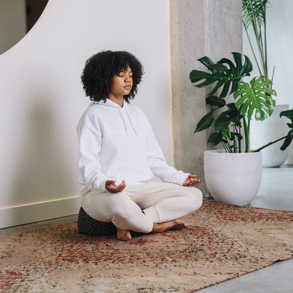 The Calm Meditation Cushion - Modern City Night photographed as a lifestyle shot with a young black woman dressed in white clothes meditating peacefully with a monstera plant in the background.  The patter on the meditation cushion is a black and white diamond pattern white spots on a background.