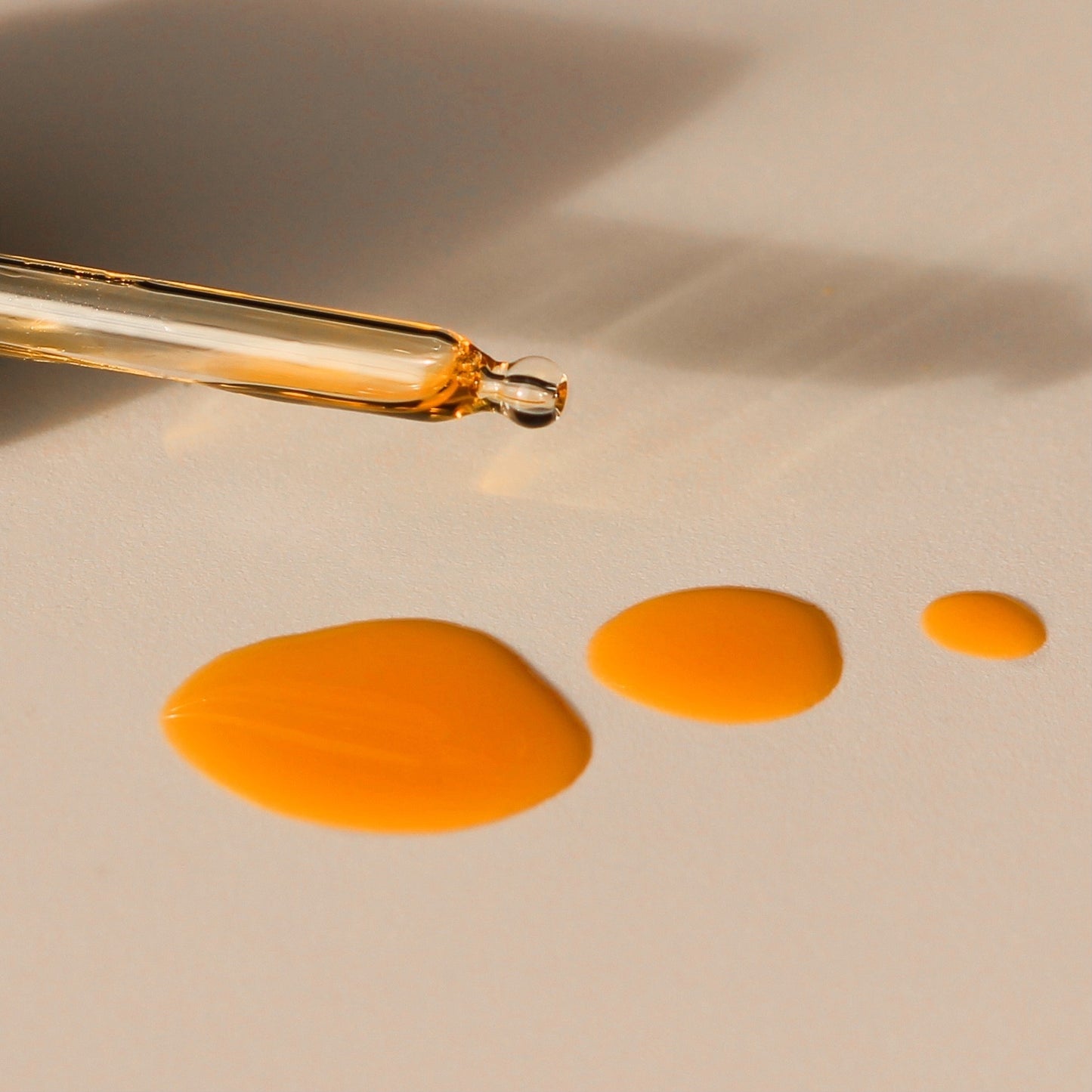 A glass dropper, dropping three golden drops of oil onto a white surface.