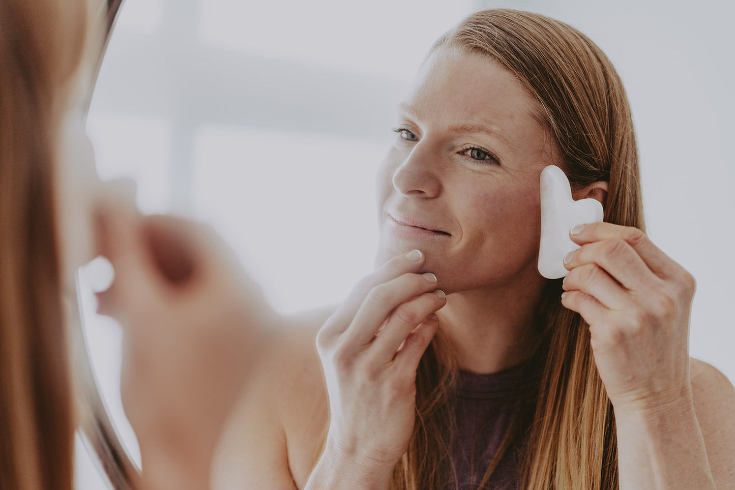 Woman looking into a mirror using a Rose Pink Gua Sha Tool on her face