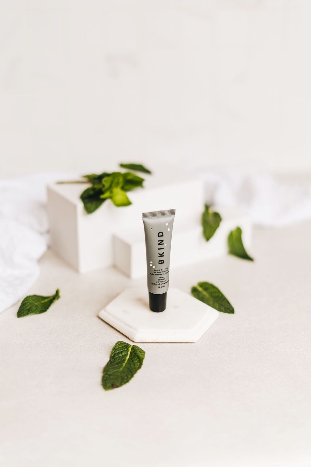 BKIND natural vegan lip balm - Peppermint - Displayed with cut peppermint leaves on three white display blocks - small grey tube of BKIND lip balm with a black lid standing upright on the white blocks.