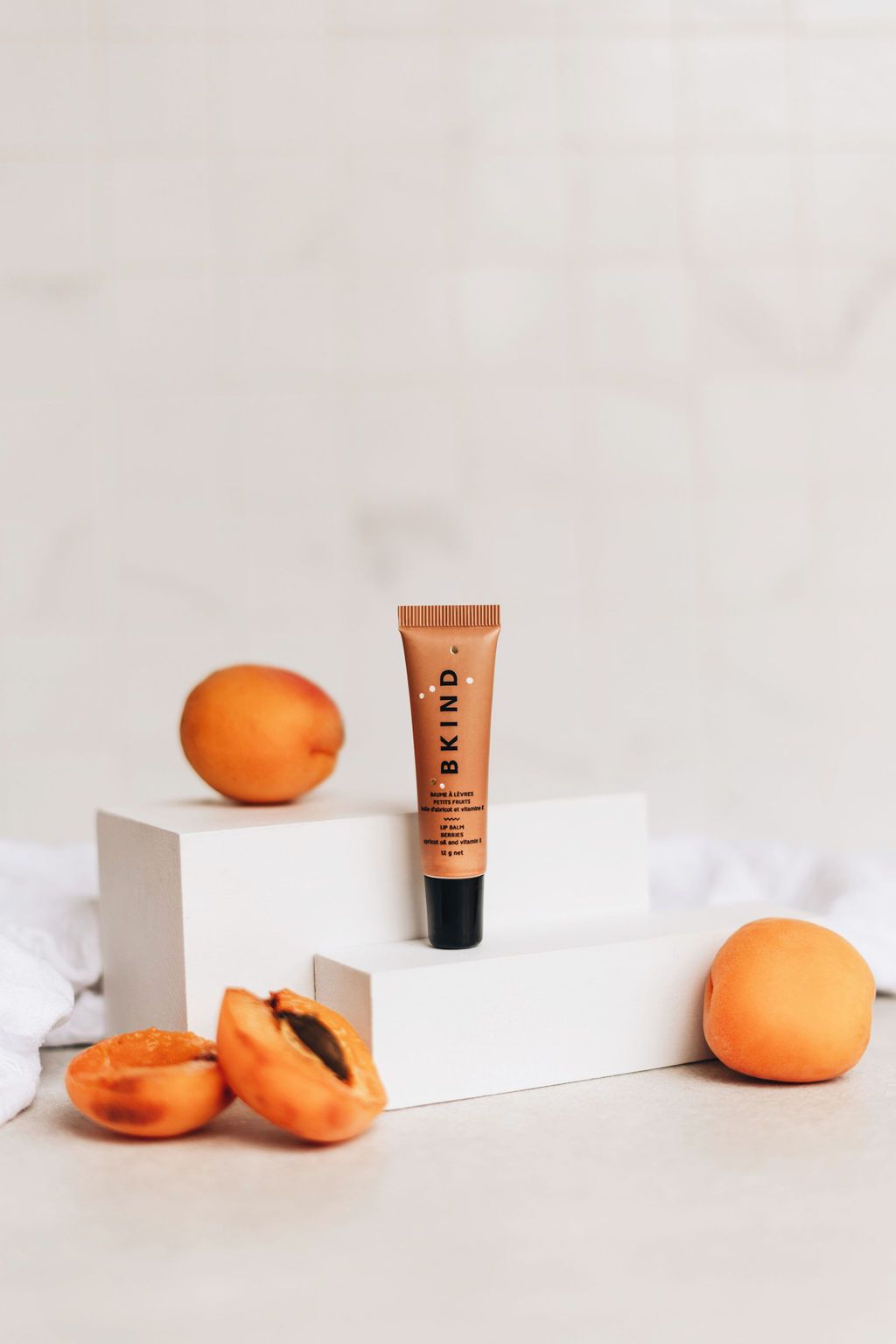 BKIND natural vegan lip balm - Berries - Displayed with full and cut apricots (the main oil used in the lip balm) on two white blocks, matching the small orange/brown tube of BKIND lip balm with a black lid standing upright on the white blocks.