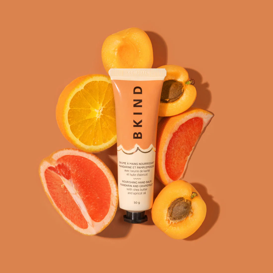 BKIND - Nourishing Hand Balm - Mandarin and Grapefruit is shown on a orange/brown background lying amongst fresh cute fruit of apricot, orange and grapefruit.