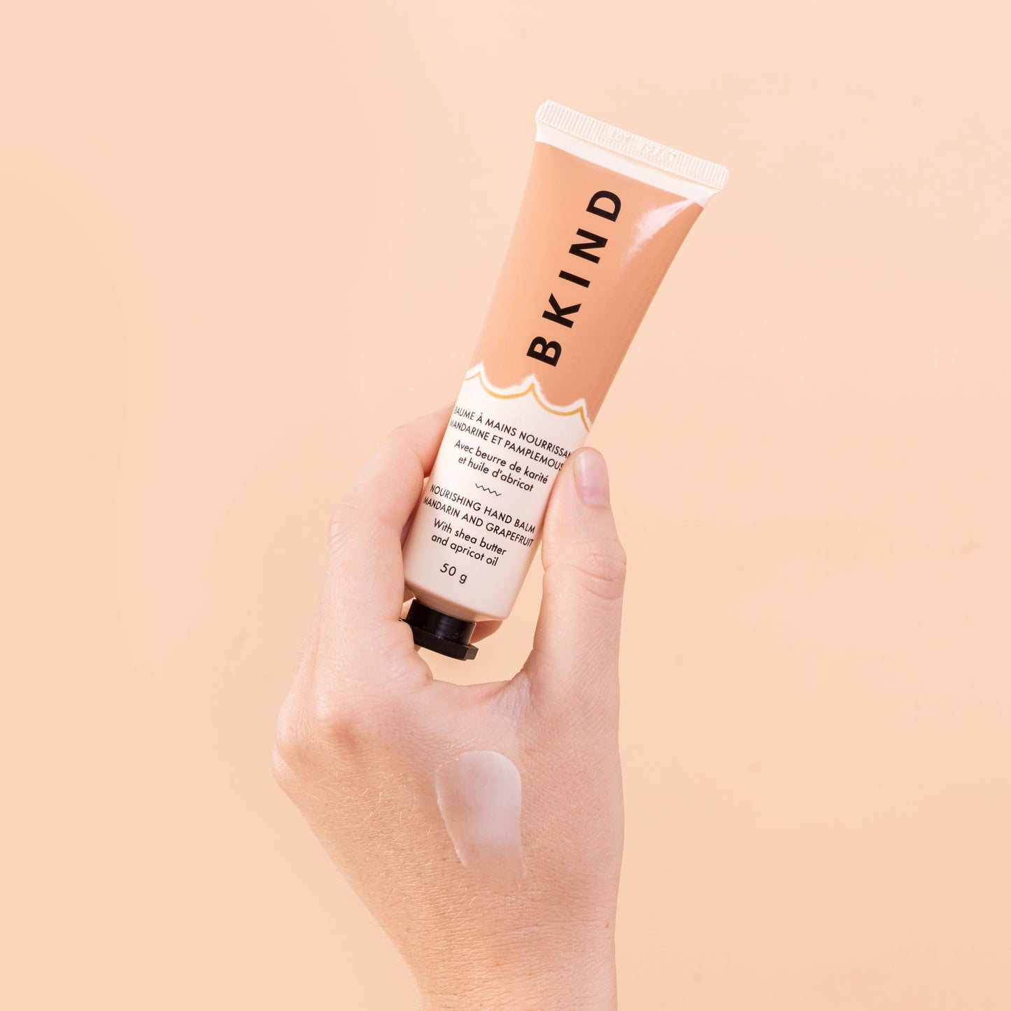 BKIND - Nourishing hand balm - Mandarin and Grapefruit - this tube is held by someones hand and shown close up - well lit on a light peach background with vertical writing of BKIND on the product label.