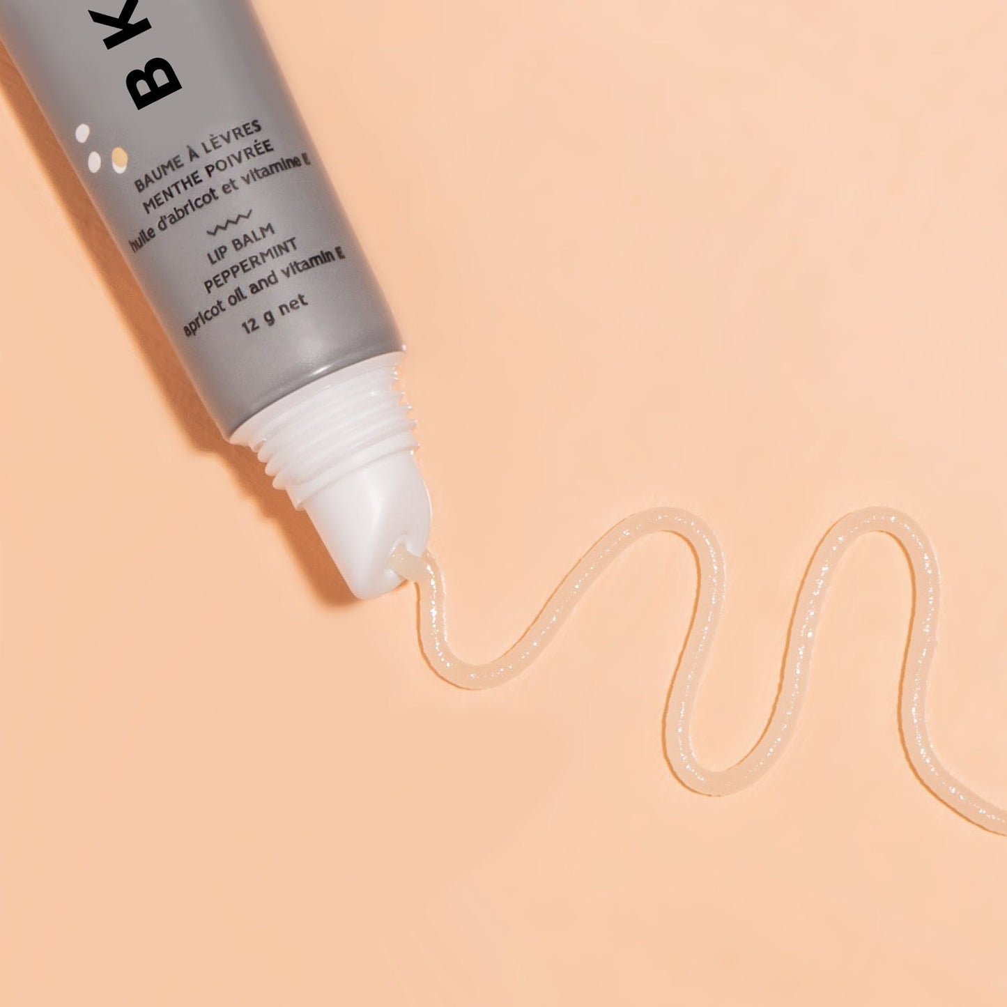 Close up of BKIND - Peppermint Lip Balm - squeezed out of grey tube onto a peach background displaying the glossy product.