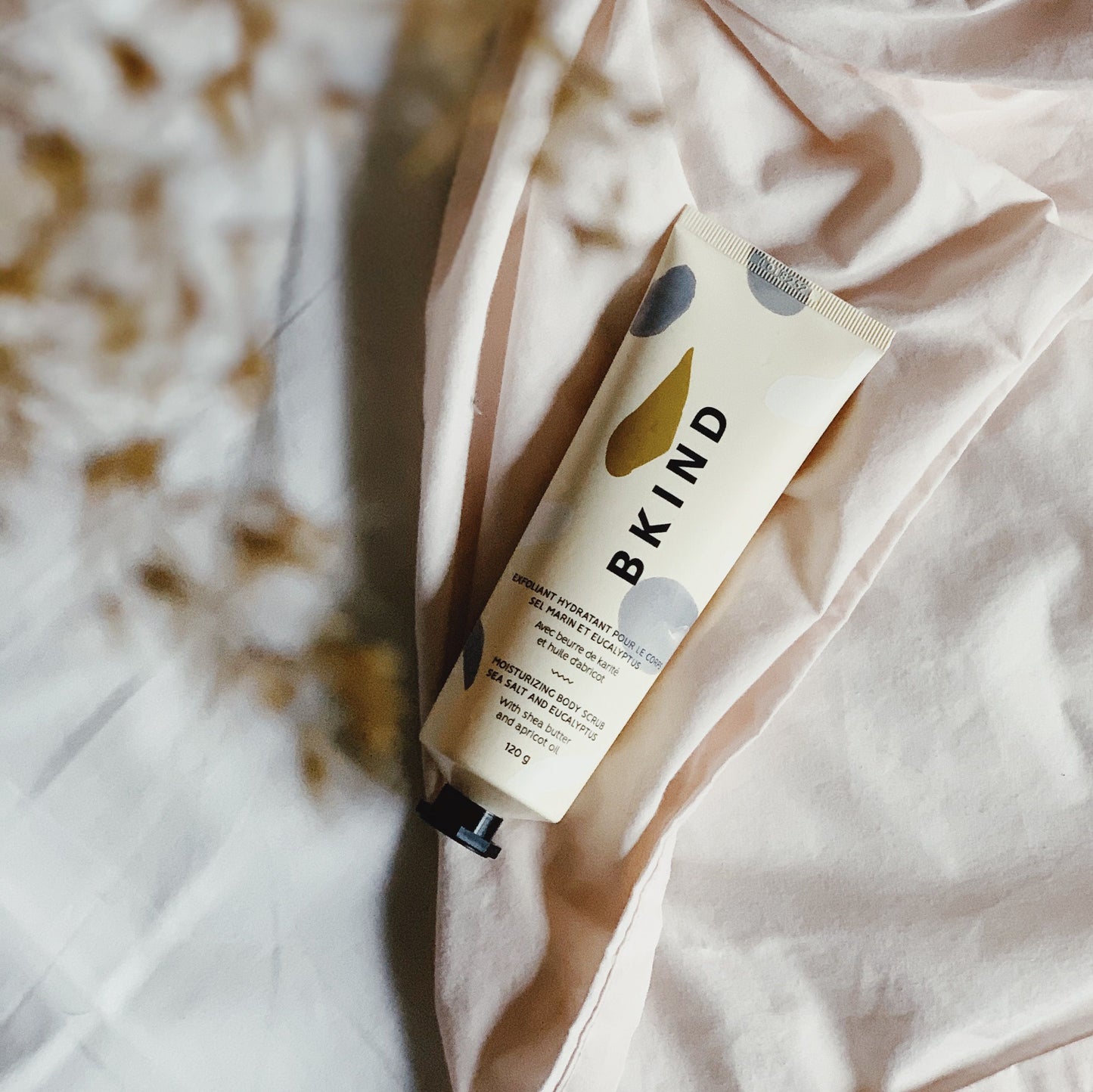 Lifestyle photo of BKIND sea salt and eucalyptus moisturizing scrub in a beige tube with powder blue designs on the tube - displayed on a cloth background with low lighting..