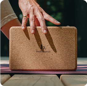 
                  
                    Close up of a hand ready to grab a cork block that is on a purple yoga mat. Nature background and bright lighting.
                  
                