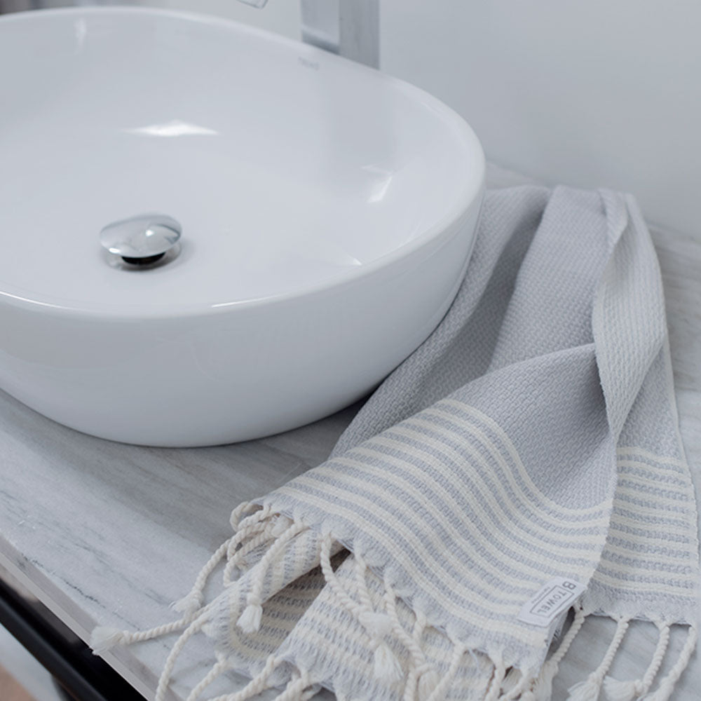 
                  
                    Lifestyle shot - Stone Grey - hand and face towel by B Yoga - Turkish Cotton laying beside tap of a white sink
                  
                