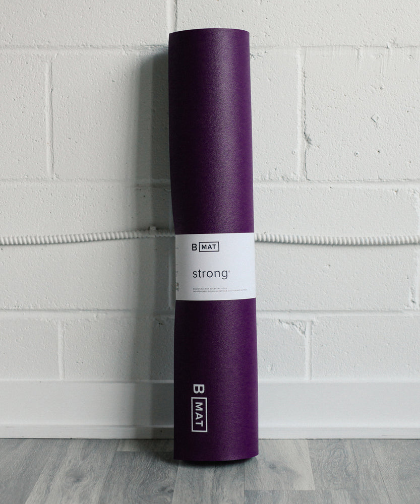 
                  
                    B Yoga - Strong 6mm yoga mat in deep purple leaning against a white brick wall
                  
                