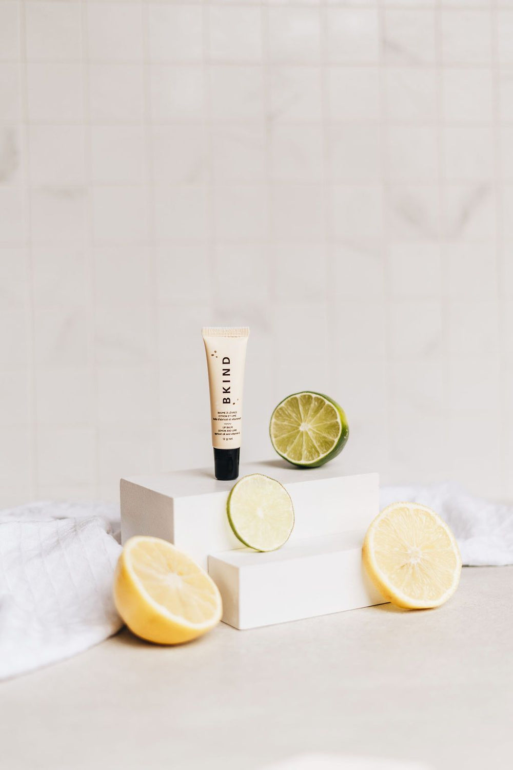 BKIND natural vegan lip balm - Lemon and Lime - Displayed with cut lemons and limes on two white blocks - small tube of BKIND lip balm with a black lid standing upright on the white blocks.