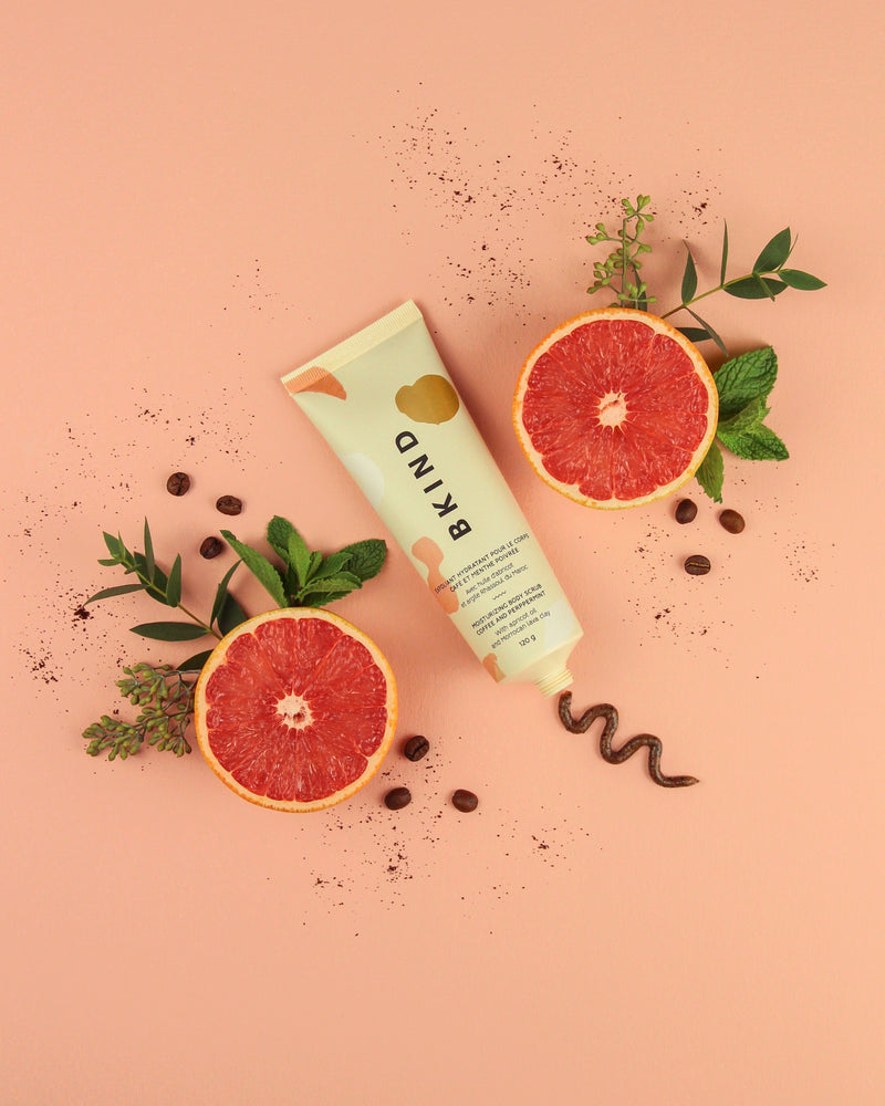 BKIND coffee and peppermint moisturizing scrub in a beige tube with peach designs on the tube - displayed beside grapefruit and fresh leaves and flowers on a peach background with a display of the coffee scrub on the background.