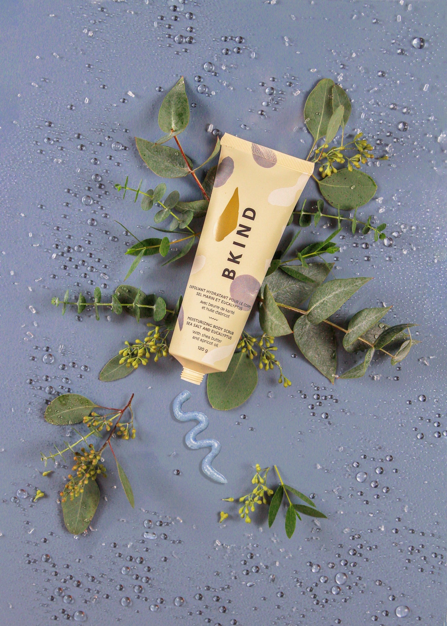 BKIND sea salt and eucalyptus moisturizing scrub in a beige tube with powder blue designs on the tube -  displayed on a blue background lying over green eucalyptus leaves with a display of the scrub on the blue background.